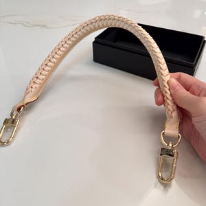 May include: A cream-colored, braided leather handbag handle with gold-tone clasps. The handle is curved and features a woven design. The clasps are attached to the handle with small, gold-colored rings. A black box is in the background.
