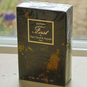 May include: A rectangular black perfume box with gold mottled accents, wrapped in clear plastic. The label reads "parfum First de Van Cleef & Arpels Paris" in gold lettering. The bottom front displays the volume as 15 ml - 0.5 FL. OZ.