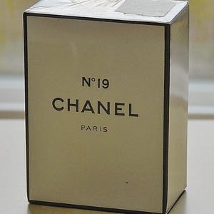 May include: A rectangular box of Chanel No. 19 perfume. The box is off-white with black text that reads "N&deg;19 CHANEL PARIS". The box has a black border and is wrapped in clear plastic.