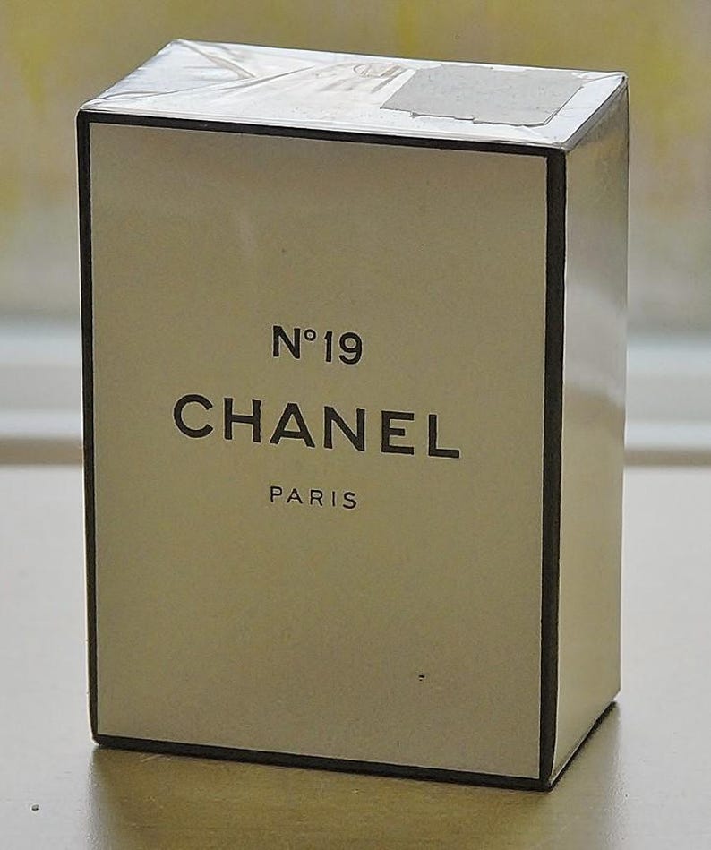 May include: A rectangular box of Chanel No. 19 perfume. The box is off-white with black text that reads "N&deg;19 CHANEL PARIS". The box has a black border and is wrapped in clear plastic.