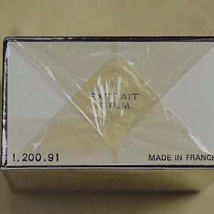 May include: A rectangular box wrapped in clear plastic. The box has white panels and black trim. Text on the box includes "EXTRAIT P.M.", "1.200.91", and "MADE IN FRANCE".