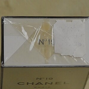May include: A close-up of a perfume box with the text "N&deg;19 CHANEL PARIS". The box is wrapped in clear plastic. The top of the box is white with the number "19" visible. The bottom of the box is gold.