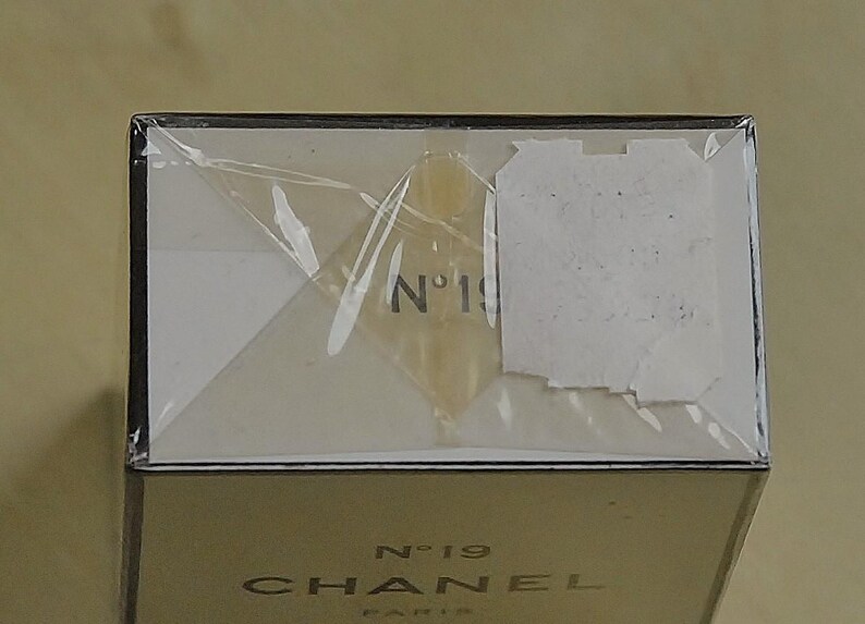May include: A close-up of a perfume box with the text "N&deg;19 CHANEL PARIS". The box is wrapped in clear plastic. The top of the box is white with the number "19" visible. The bottom of the box is gold.