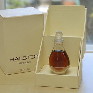 May include: A teardrop-shaped glass perfume bottle with a clear stopper, containing amber liquid. The bottle is labeled Halston and sits in an open cream-colored box. The box exterior displays the text Halston Perfume 1/2 fl. oz.