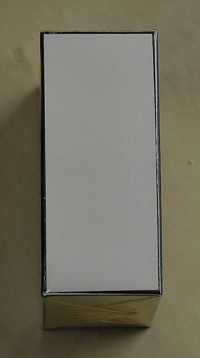 May include: A rectangular box with a white top and gold bottom. The edges are outlined in black. The box is sitting on a light brown surface.