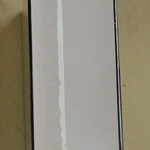 May include: A rectangular product box with a white front and a gold-colored bottom. The box is wrapped in clear plastic. The edges of the box are black. The box is sitting on a light brown surface.