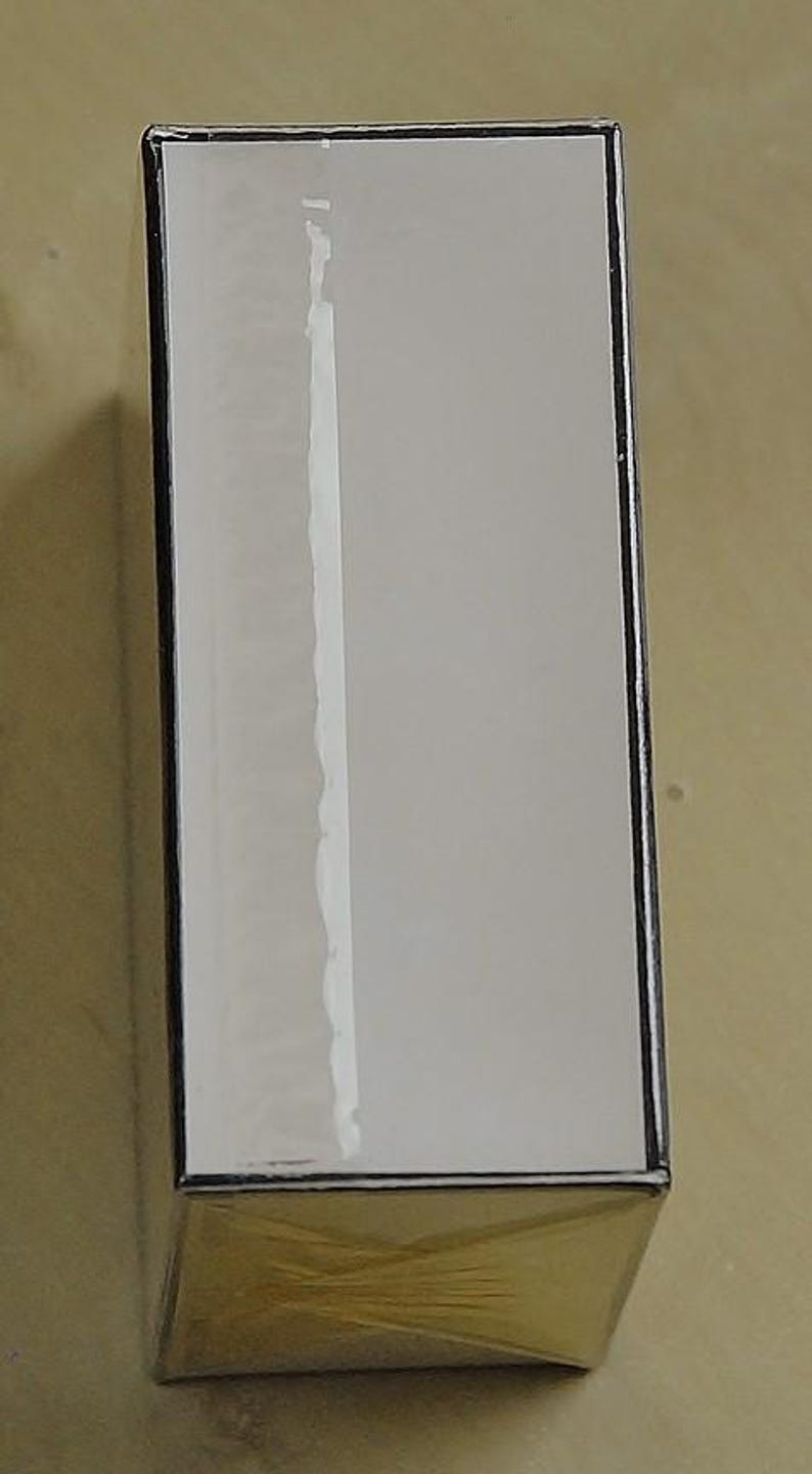 May include: A rectangular product box with a white front and a gold-colored bottom. The box is wrapped in clear plastic. The edges of the box are black. The box is sitting on a light brown surface.
