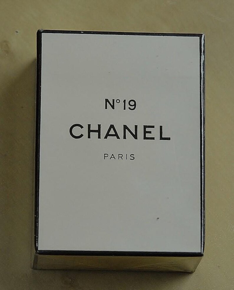 May include: A rectangular box with a white front and gold sides. The front of the box features the text "N&deg;19 CHANEL PARIS" in black, centered on the white background. The box has a black border.
