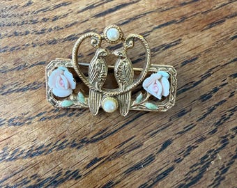 Op de afbeelding: Goudkleurige broche met twee vogels die elkaar aankijken, met een hartvormig ontwerp erboven. De broche bevat twee roze rozen en een parelachtig accent. Het ontwerp is geplaatst op een rechthoekige basis.