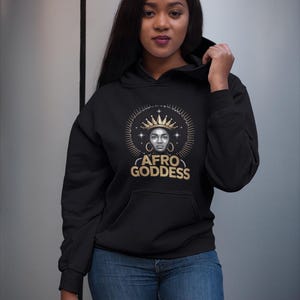 Può includere: Felpa nera con grafica dorata di una donna con corona, orecchini e la scritta "AFRO GODDESS". La felpa ha una tasca frontale e viene abbinata a jeans blu. La donna tiene su il cappuccio.