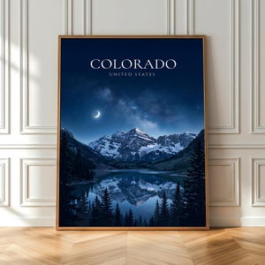 May include: A framed print featuring a nighttime landscape of Colorado, United States. The image showcases snow-capped mountains reflected in a lake under a starry sky and crescent moon. The word "COLORADO" is displayed at the top.