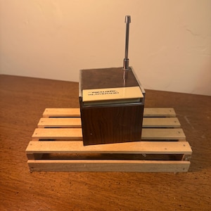 Peut inclure: Une radio Realistic Weatheradio vintage, avec une finition en imitation bois foncé et une antenne argentée. La radio est posée sur une petite palette en bois. Le texte "REALISTIC WEATHER RADIO" est visible à l'avant.