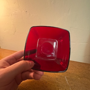 Puede incluir: Un cenicero cuadrado de vidrio rojo rubí con un borde negro. El cenicero tiene una hendidura circular en el centro y está sostenido en la mano de una persona. El cenicero está sobre una superficie de madera.