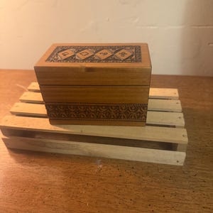 Puede incluir: Una pequeña caja de madera rectangular con intrincados diseños tallados en la tapa y la sección inferior. La caja es de color marrón cálido y se asienta sobre un palé de madera en miniatura. Los grabados presentan patrones geométricos y florales.