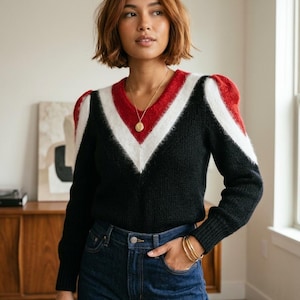 May include: A black V-neck jumper with red and white detailing on the shoulders and neckline. The model is wearing dark blue jeans and gold jewellery. The jumper has long sleeves. The style is casual and chic.
