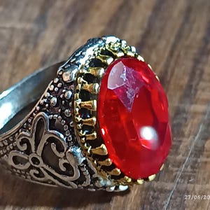 Può includere: Un anello d'argento con un design ornato e una grande pietra preziosa rossa di forma ovale. La gemma è il punto focale, incastonata in una lunetta color oro. La fascia dell'anello presenta volute dettagliate.