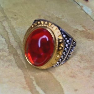 Può includere: Un anello con una grande pietra rossa traslucida di forma ovale incastonata in una montatura color oro. La fascia è realizzata in metallo color argento con dettagli scuri e strutturati sui lati, che contrastano con la montatura liscia e lucida color oro che circonda la gemma rossa centrale.