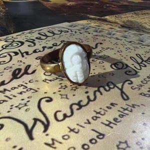 May include: A gold-toned ring with an oval, white cameo featuring a carved face. The ring rests on a surface with handwritten text including the word "Waxing" and phrases like "Healing" and "Success."
