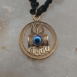 May include: Gold-colored pendant necklace with a circular design. The pendant features a trident, the text "महादेव", and a blue eye. The necklace has a black cord.