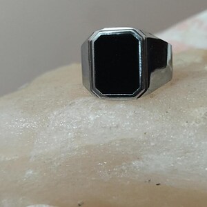 May include: A silver-toned ring with a rectangular black stone. The ring has a classic design with a polished finish. The black stone is set within a geometric bezel, creating a bold and elegant look. This ring is suitable for everyday wear.