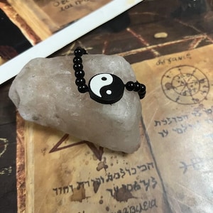 May include: A black beaded bracelet with a white and black yin yang symbol. The bracelet is resting on a light-colored, rough-textured stone. The background features aged paper with script.