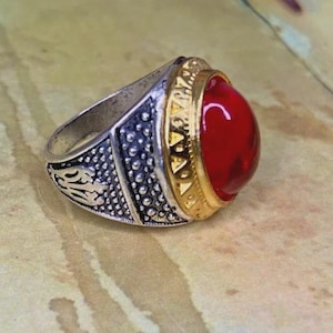 Op de afbeelding: Een zilverkleurige ring met een grote, gladde, ronde rode steen in een goudkleurige zetting. De ringband heeft een textuurrijke, geoxideerde zilveren afwerking met ingewikkelde reliëfpatronen en een kralenrand rondom de centrale rode steen.