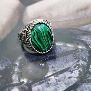 Könnte beinhalten: Ein silberfarbener Ring mit einem ovalen, grünen Malachitstein mit schwarzen Wirbelmustern. Der Stein ist in eine gedrehte Kordelbordüre gefasst, und die Ringschiene weist an den Seiten ein strukturiertes, filigranes Metalldesign auf.