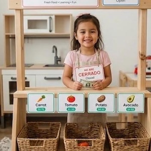Op de afbeelding: Een houten marktkraam met borden in het Engels en Spaans. De borden luiden "Welcome to the Little Market" en "Open". Hieronder staan borden voor wortel, tomaat, aardappel en avocado. Een jong meisje staat achter de kraam.