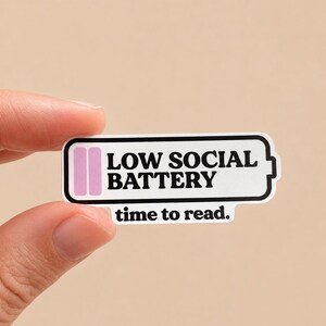 May include: A rectangular sticker with rounded corners featuring a battery icon with two pink bars. The text reads LOW SOCIAL BATTERY in bold black capital letters, followed by the text time to read. in smaller black lowercase letters.
