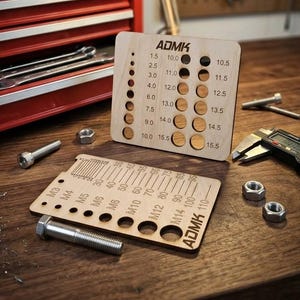 May include: Wooden bolt and nut size gauges with the text "ADMK" and measurement markings. A red toolbox, a desk lamp, and various tools are in the background. The gauges are on a wooden surface.