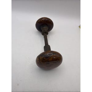 Puede incluir: Un juego de pomos para puerta que consta de dos pomos redondos de mármol marrón y bronceado, conectados por un eje central de metal. Los pomos tienen un acabado liso y brillante con patrones en espiral, y la varilla de conexión es de metal gris oscuro.