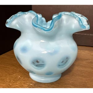 May include: A light blue hobnail glass vase with a ruffled, crimped rim. The body of the vase features a repeating pattern of raised, rounded bumps, and the edges of the ruffled top are accented with a slightly darker shade of blue glass.