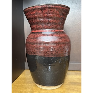 May include: A ceramic vase featuring a two-tone glaze design. The upper portion displays a textured, mottled red and black finish with horizontal ridges, while the lower section has a smooth, glossy solid black glaze and a light beige base.