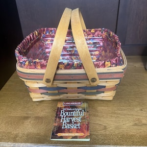 Puede incluir: Una cesta tejida rectangular con dos asas de madera, con bandas horizontales de madera natural, azul oscuro y rojo apagado. El interior está forrado con una tela con volantes y un patrón de hojas de otoño. Una tarjeta de producto dice Longaberger 1997 Shades of Autumn Bountiful Harvest Basket.