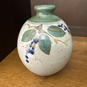 May include: A ceramic vase with a round body and a narrow neck. The vase is off-white with a speckled texture and is decorated with green leaves and blue grape clusters. The rim and neck are green.