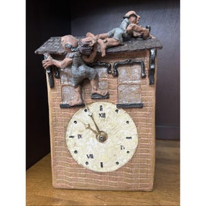 May include: A rectangular clay clock with a textured brick-patterned surface. Two sculpted figures in grey textured clothing are positioned on the roof and side. The clock face is circular, cream-colored, and features black Roman numerals and hands.