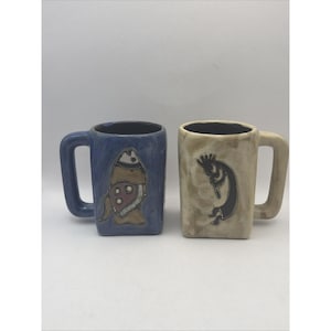 Op de afbeelding: Twee keramische mokken met vierkante lichamen en hoekige handvatten. De linker mok is blauw met een bruine en witte visillustratie. De rechter mok is beige met een donkerbruine illustratie van een fluitspelende figuur.