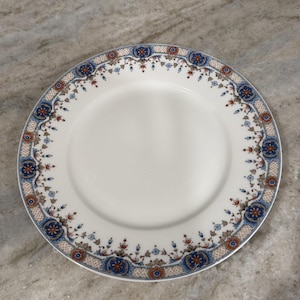 May include: A round white ceramic dinner plate featuring a decorative border pattern. The rim is adorned with alternating blue and rust-colored floral medallions connected by intricate geometric lattice bands and small blue floral sprigs.