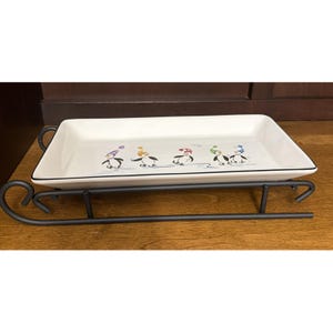 May include: A rectangular white ceramic serving tray with a black rim, featuring a graphic of five penguins wearing colourful winter hats and scarves. The tray is supported by a black metal stand shaped like a traditional sled with curved runners.