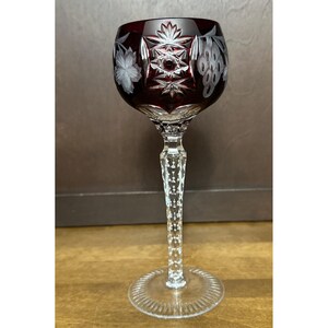 May include: A deep red cut-to-clear crystal wine glass featuring intricate etched star and grape cluster patterns on the bowl. The stem is faceted with a diamond-cut texture, and the circular base has a radial ribbed design around the edge.