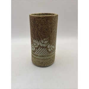 May include: A cylindrical ceramic tumbler with a speckled brown and tan finish. The exterior features a carved band around the lower section displaying a repeating pattern of stylized leaves and vertical lines in a lighter, off-white clay tone.