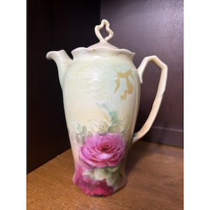 Puede incluir: Una cafetera de porcelana con fondo amarillo pálido y crema, decorada con una gran rosa de color rosa intenso en la base. La pieza presenta un borde festoneado, un asa curva, un pico vertedor y una tapa a juego con un remate decorativo.
