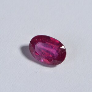 May include: A vibrant, oval-shaped ruby gemstone. The gem displays a deep red hue with a polished surface, set against a white background. The ruby's elongated form and rich color are prominent.