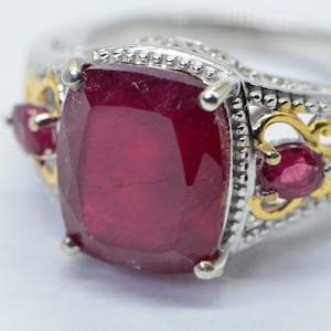 May include: A silver and gold ring featuring a large, square-cut ruby gemstone as the centerpiece. The ring is accented with smaller ruby stones and ornate gold filigree detailing. The band has a silver finish.