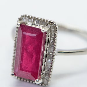 May include: A silver ring featuring a large, rectangular, deep red gemstone. The gemstone is surrounded by a halo of smaller, clear stones. The ring's band is thin and polished, with a bright, reflective surface. The setting is detailed and elegant.