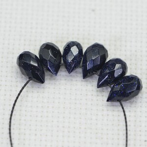 May include: A strand of dark blue faceted gemstone beads, strung on a black thread. The beads are teardrop shaped and have a sparkly, speckled finish.