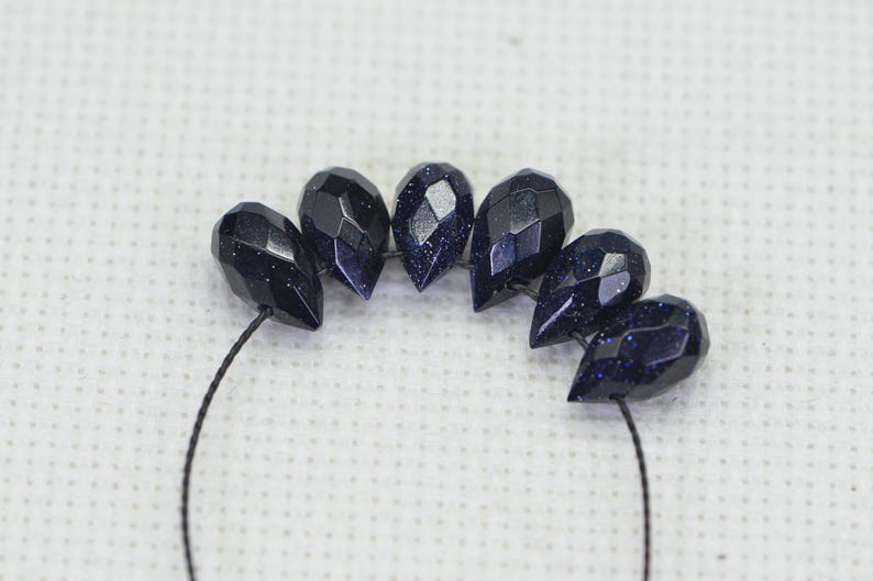 May include: A strand of dark blue faceted gemstone beads, strung on a black thread. The beads are teardrop shaped and have a sparkly, speckled finish.