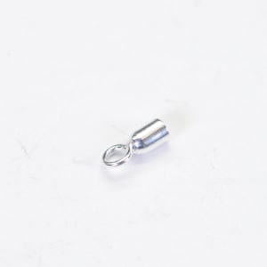 May include: A small, silver-colored jewelry finding, likely a crimp end or clasp component, lies on a white surface. The piece has a cylindrical body with a loop at one end, designed for attaching to other jewelry elements.