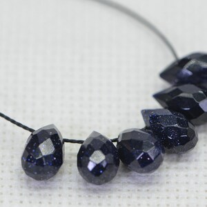 May include: A strand of dark blue gemstone beads, each bead is faceted and shaped like a teardrop. The beads are strung on a black cord.