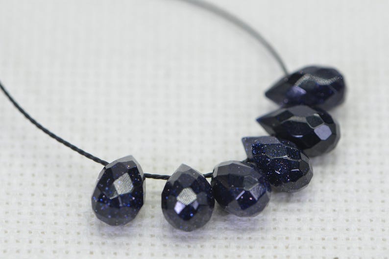 May include: A strand of dark blue gemstone beads, each bead is faceted and shaped like a teardrop. The beads are strung on a black cord.